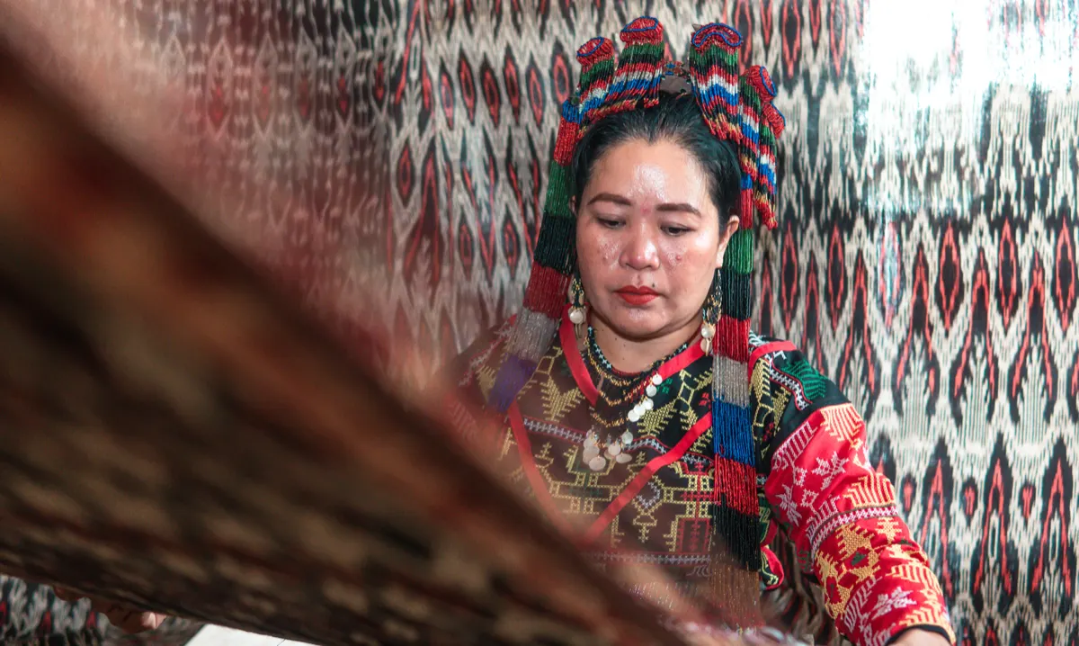 A weaver in a beaded headpiece at her loom, T’nalak hanging behind her
