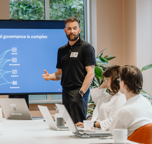 Man in black polo presenting AI governance topic to two seated colleagues with laptops in a meeting room.