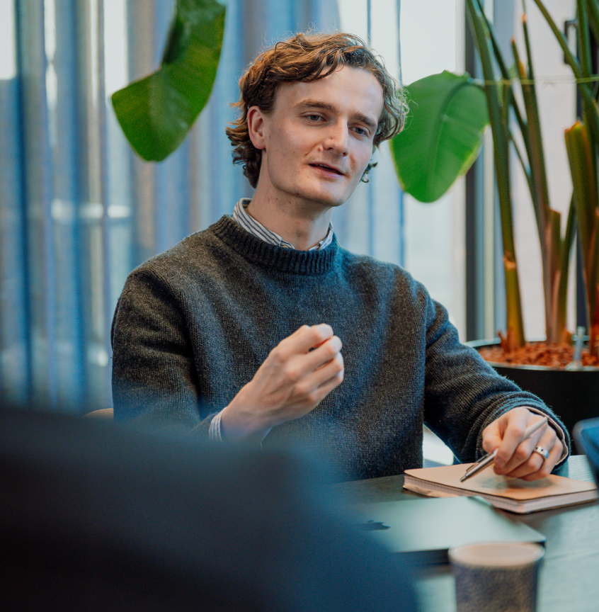 Man in gray sweater gestures while seated at table with notebook and pen near plants.