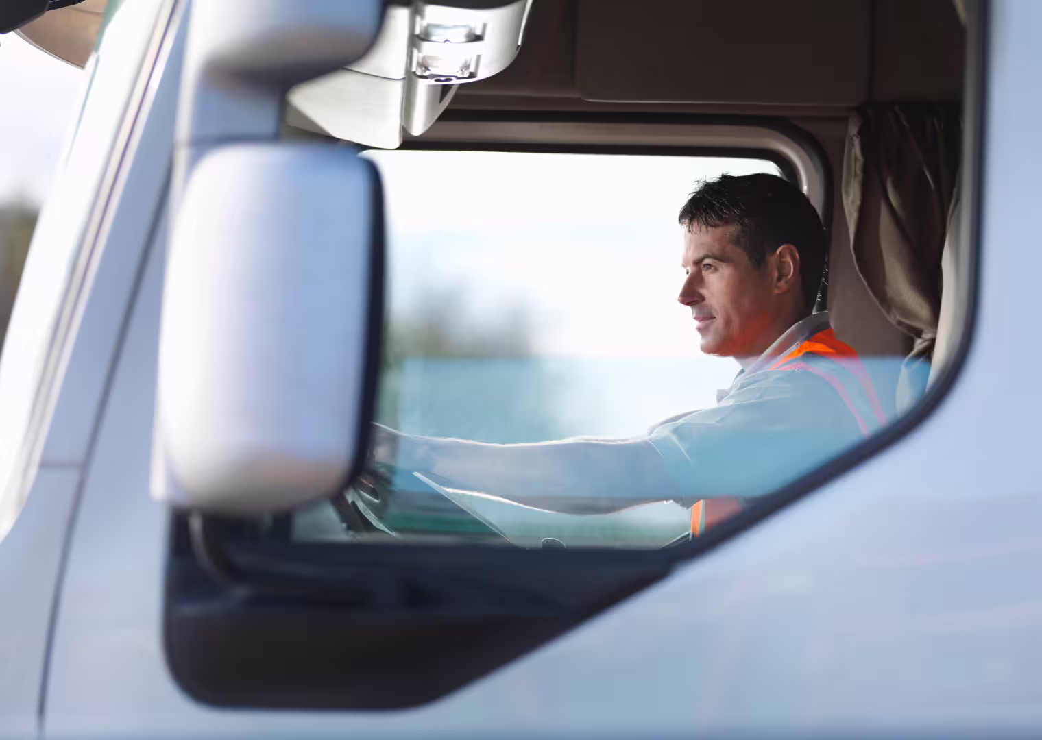 Side view of a man wearing an orange safety vest driving a truck, visible through the truck's window and side mirror.