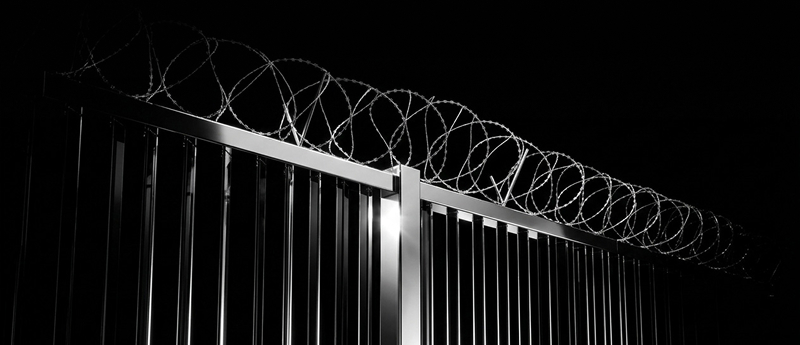Black metal fence topped with spiraled razor wire against a dark background, illuminated by a light source.