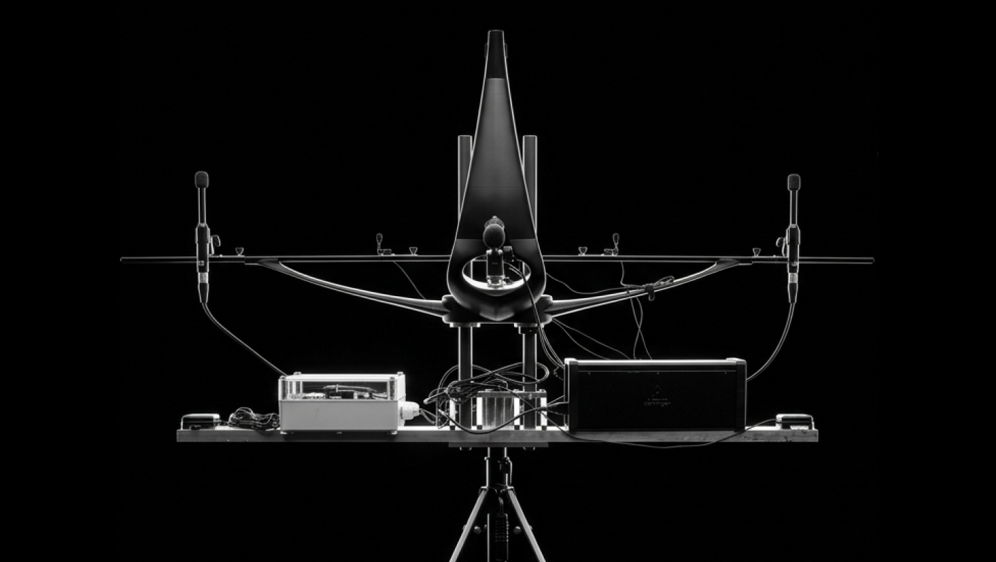 Front symmetrical view of a complex electronic setup on a tripod against black background featuring a central cone-shaped structure flanked by two microphones on stands connected by cables to electronic devices on a platform.