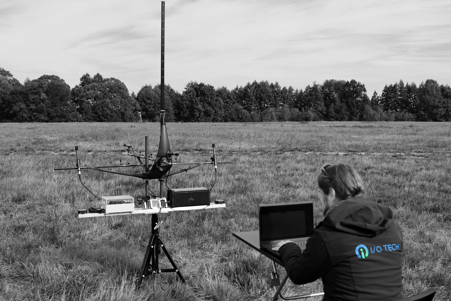 Person wearing a jacket with the 'I/O TECH' logo seated at a portable table working on a laptop in a grassy field near a tripod-mounted scientific instrument with antennas and electronic equipment, surrounded by trees in the background.