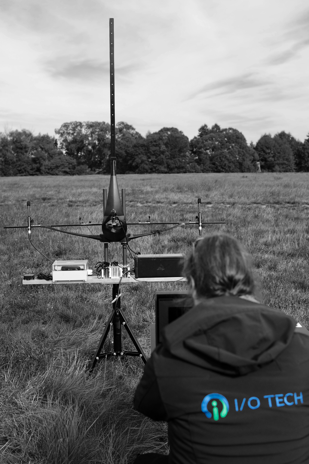Person in an I/O Tech jacket operating a laptop outdoors in a grassy field, facing a scientific instrument mounted on a tripod with antennas and electronic components, against a backdrop of trees and cloudy sky.