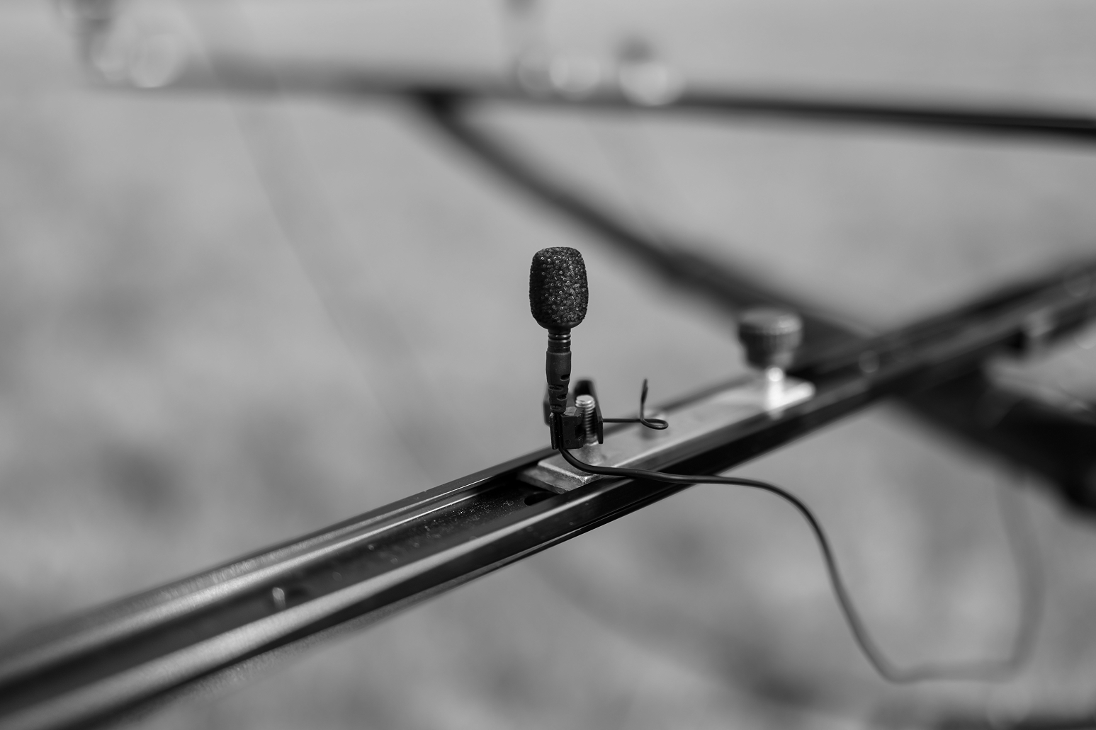 Close-up of a small microphone with a foam windscreen mounted on a metal rail, attached by a clip with a cable extending out of frame.