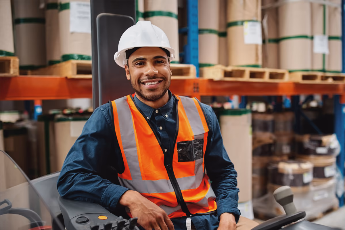 Smiling warehouse worker in a hard hat and safety vest seated at a forklift