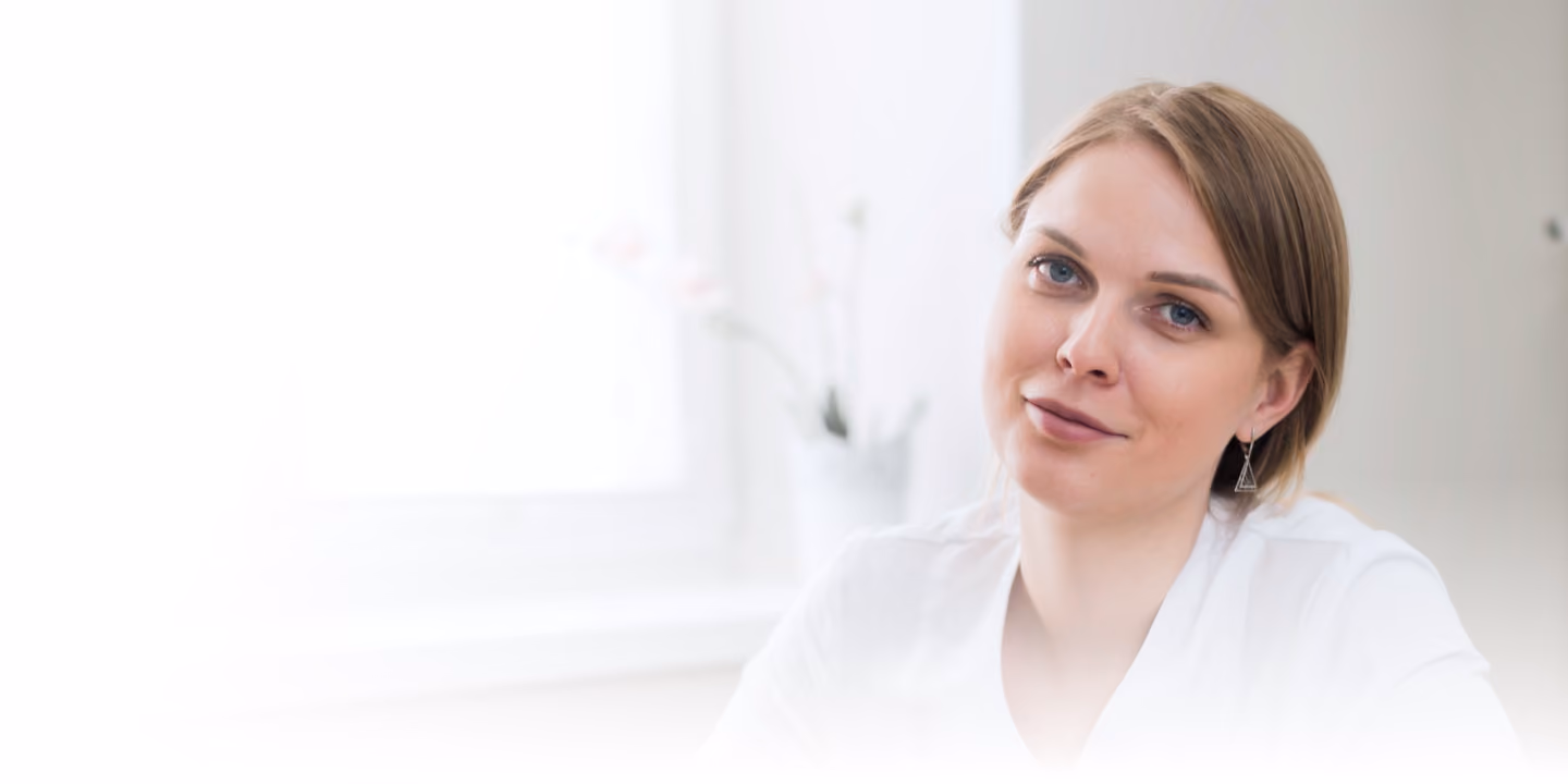 A woman with light brown hair and blue eyes wearing a white top, smiling softly in bright indoor lighting near a window.
