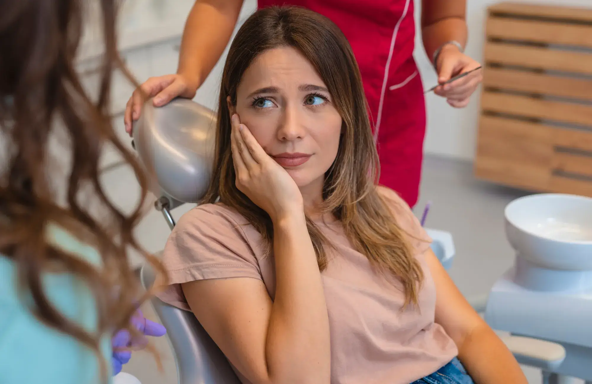 Woman in a dental chair touching her cheek with a concerned expression while talking to a dental professional.