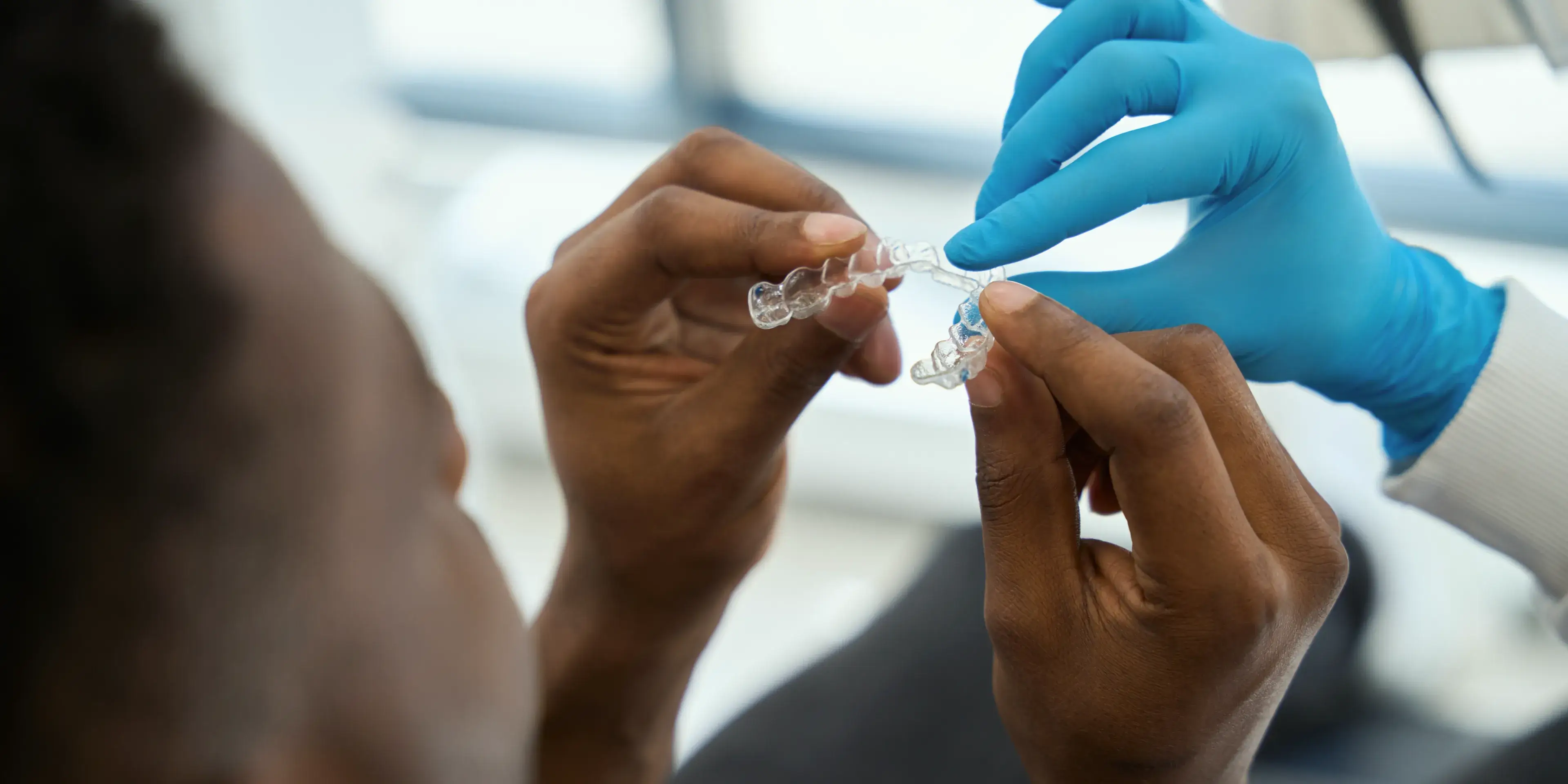A person holding a clear dental aligner while a gloved hand points at it during a dental fitting.