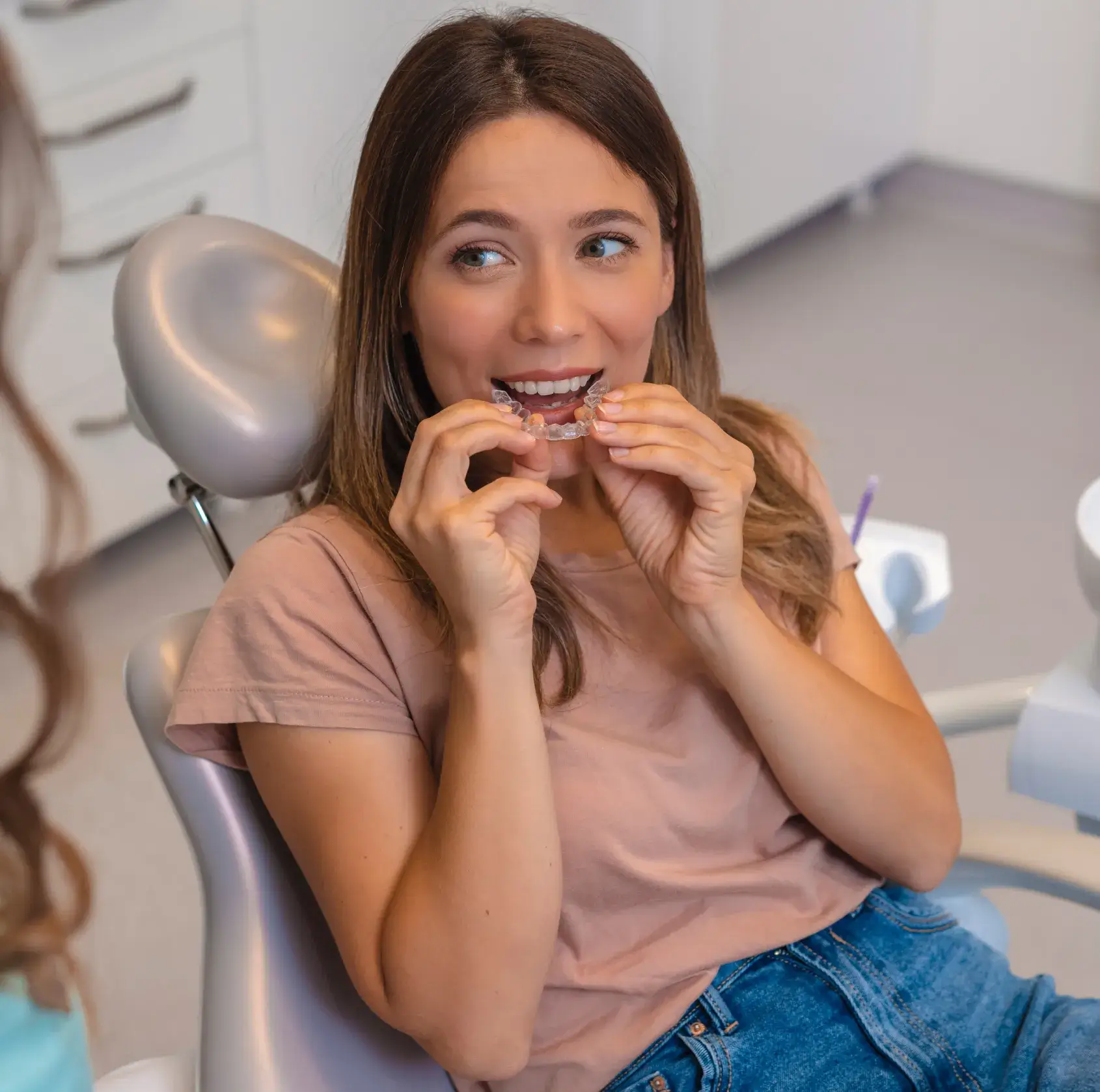 Young woman sitting in dental chair holding a clear orthodontic aligner near her mouth.