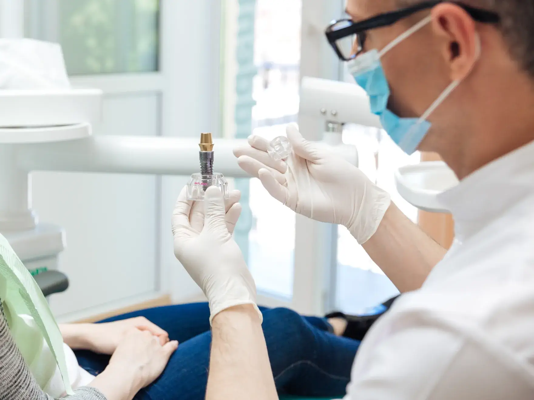 Dentist wearing gloves and mask holding a dental implant model in front of a patient seated in the chair.