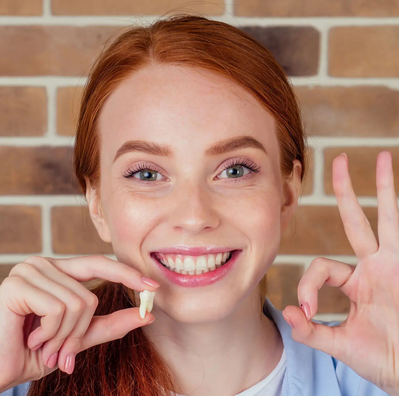 Smiling red-haired woman holding a removed tooth and making an OK hand gesture in front of a brick wall.