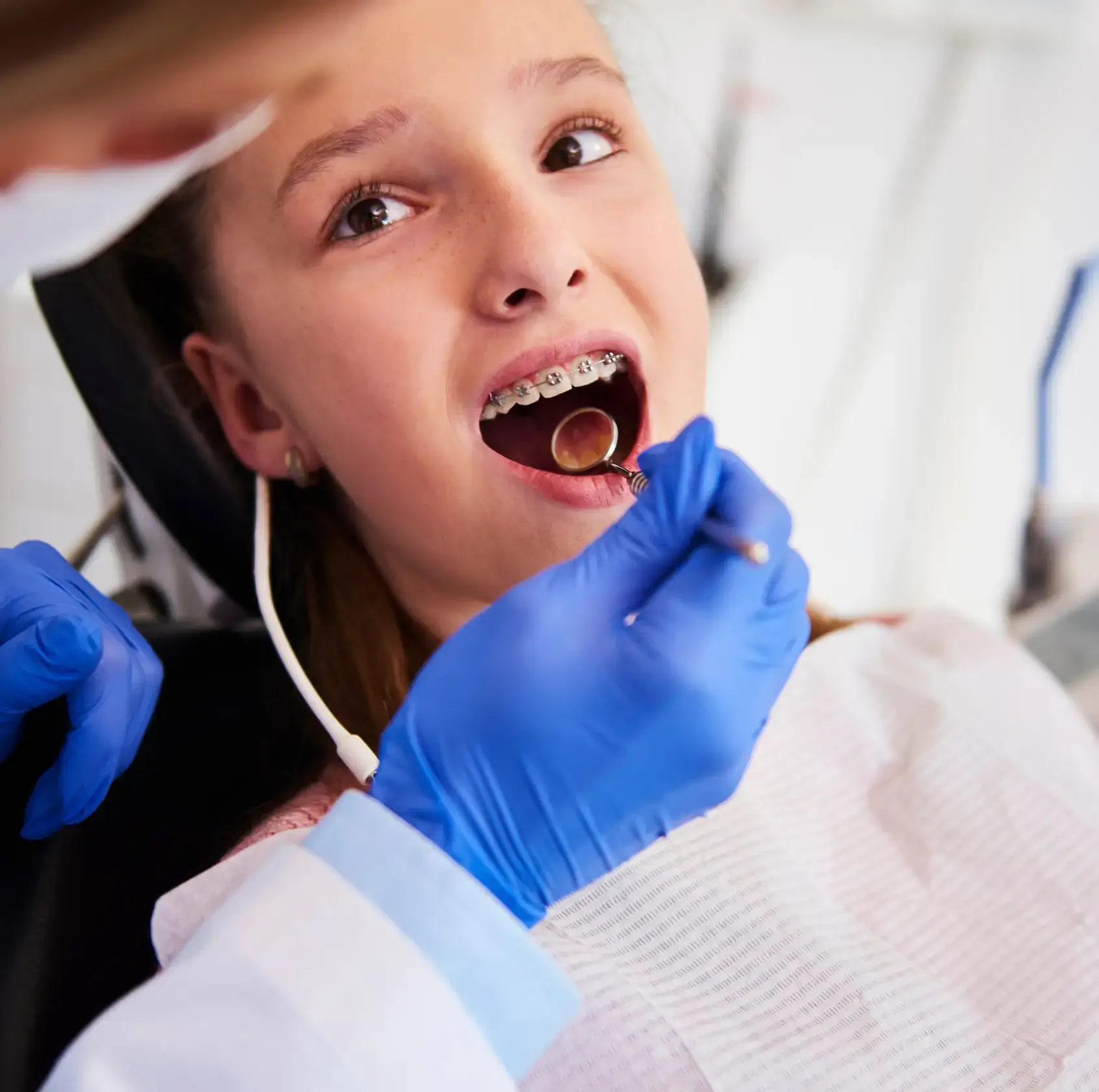 Dentist wearing blue gloves examining a girl's braces using a dental mirror.