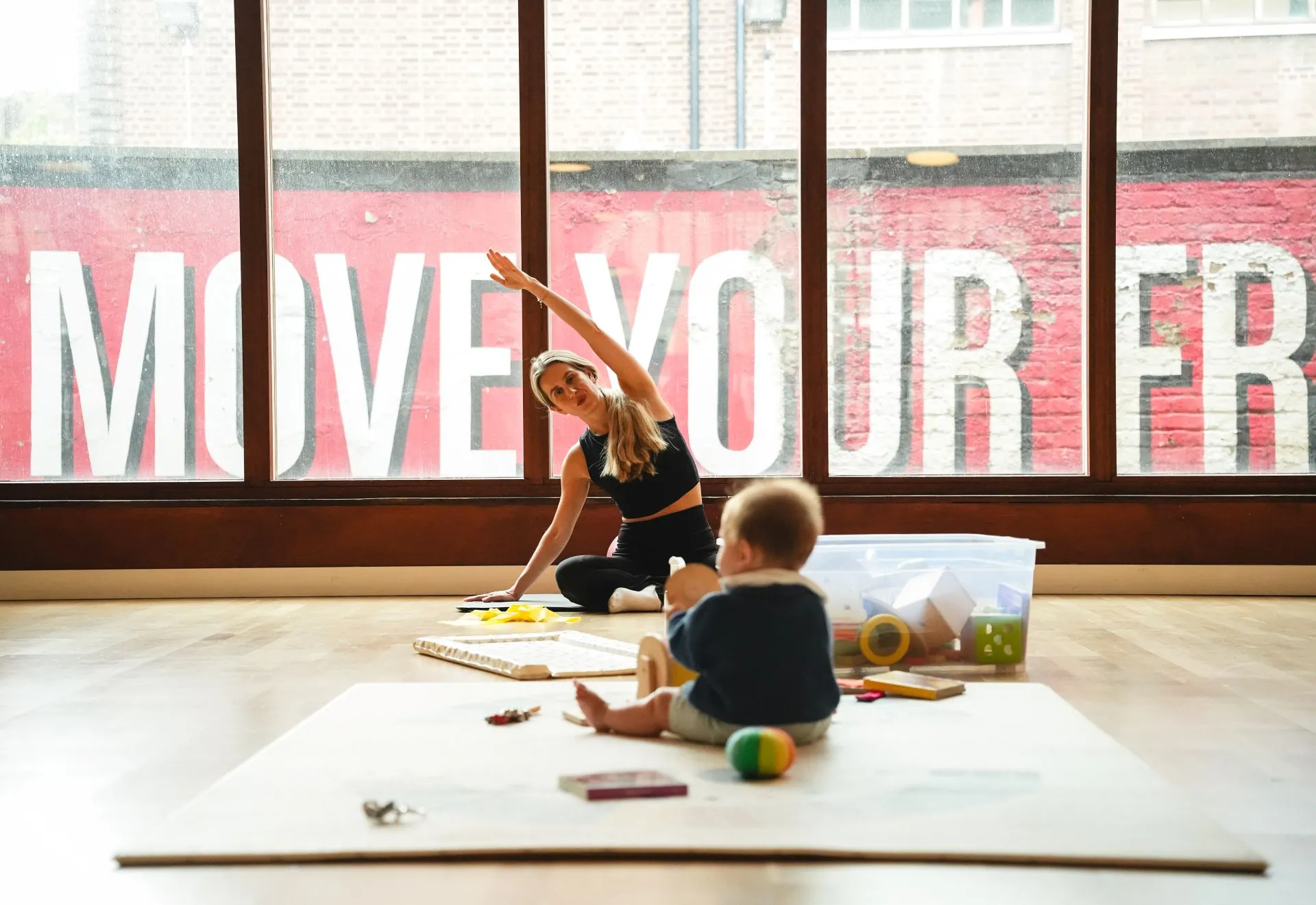 A woman in black activewear sitting on the floor stretching her arm overhead in a spacious room with large windows, while a baby sits on a mat nearby surrounded by toys.
