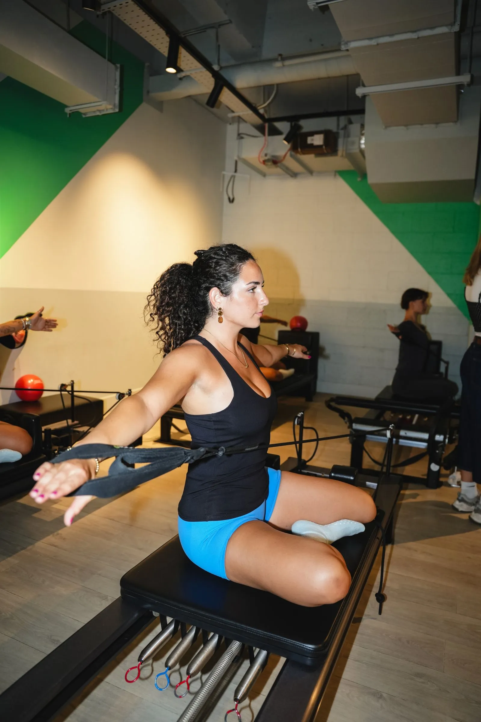 Woman with curly hair in a black tank top and blue shorts exercises on a Pilates reformer machine, holding the straps with arms extended sideways in a studio with wooden floors and green and beige walls.