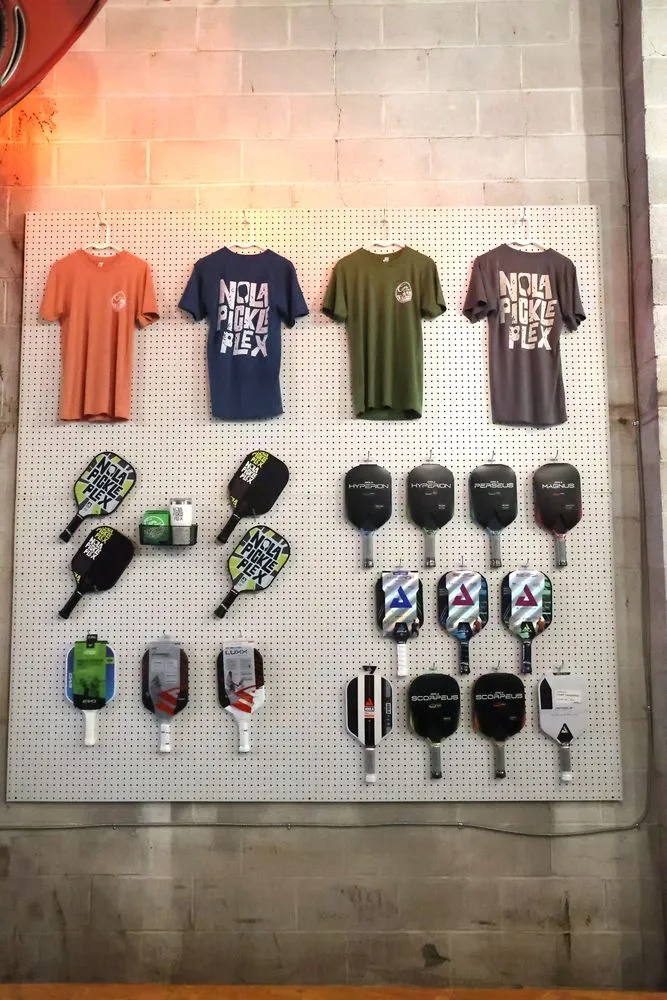 Pro shop wall with pickleball paddles and NOLA PicklePlex T-shirts