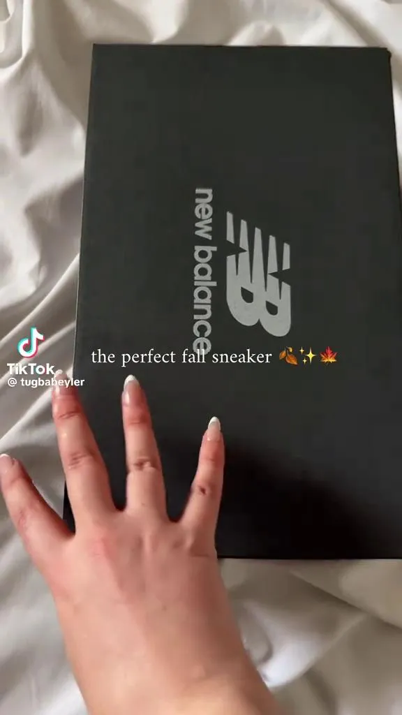 A hand reaching for a New Balance shoebox on a bed