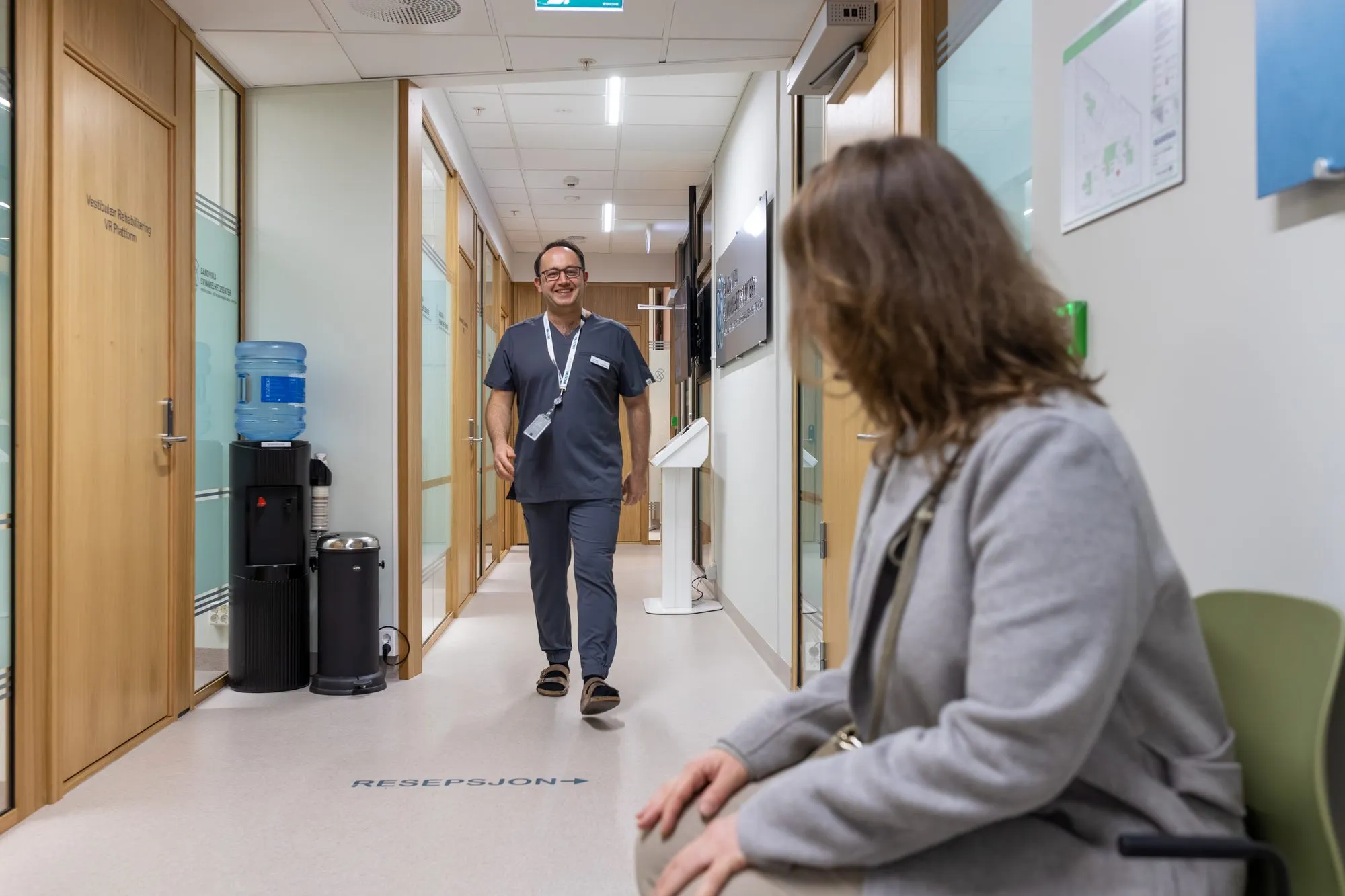 Behandler går mot pasient som venter i klinikkens korridor ved resepsjonen
