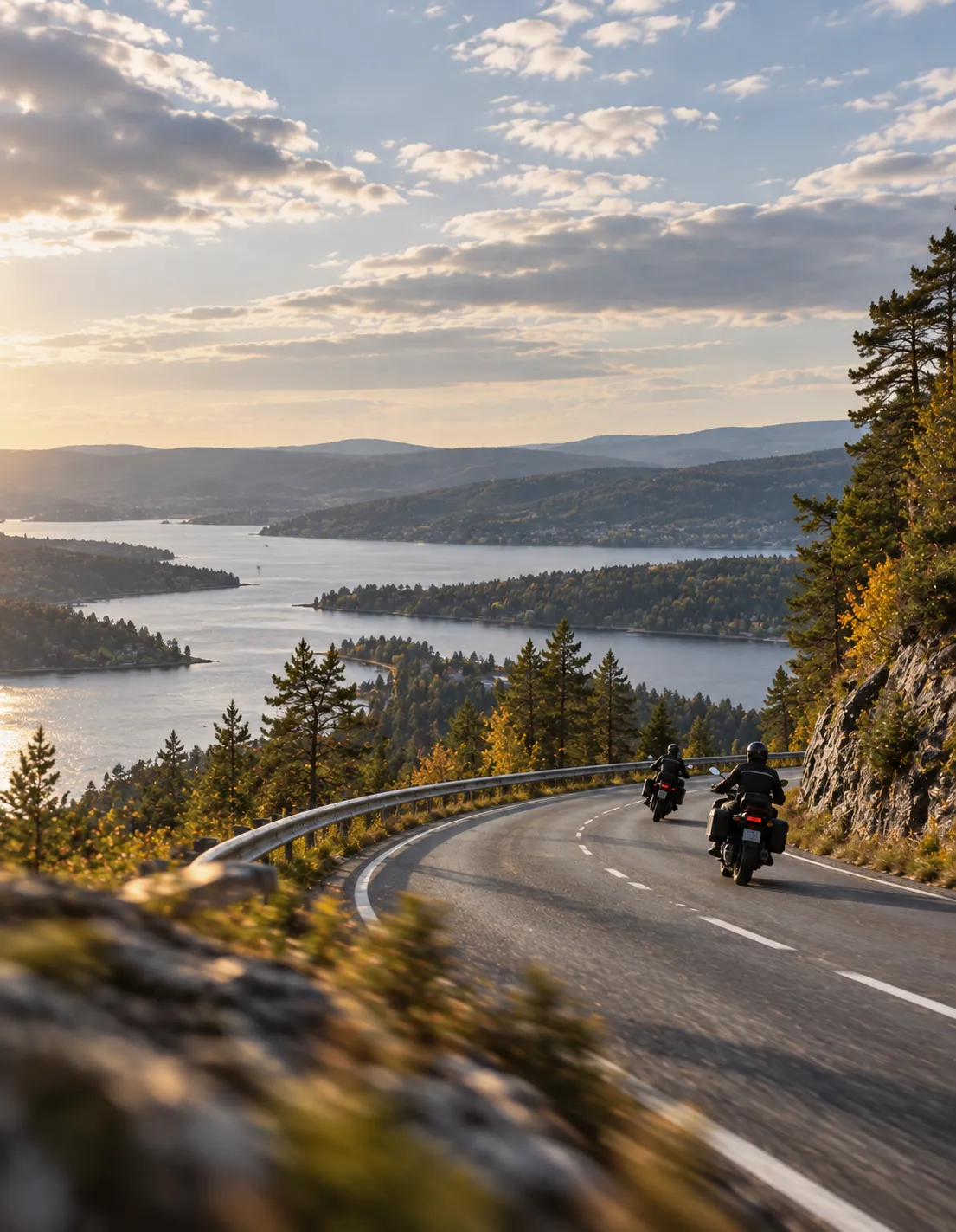To motorsykler på en svingete vei med utsikt over fjord og åser i kveldssol