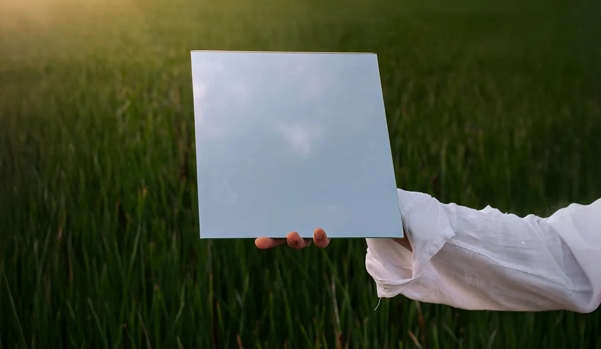 Hand holding a mirror reflecting the sky in a green field