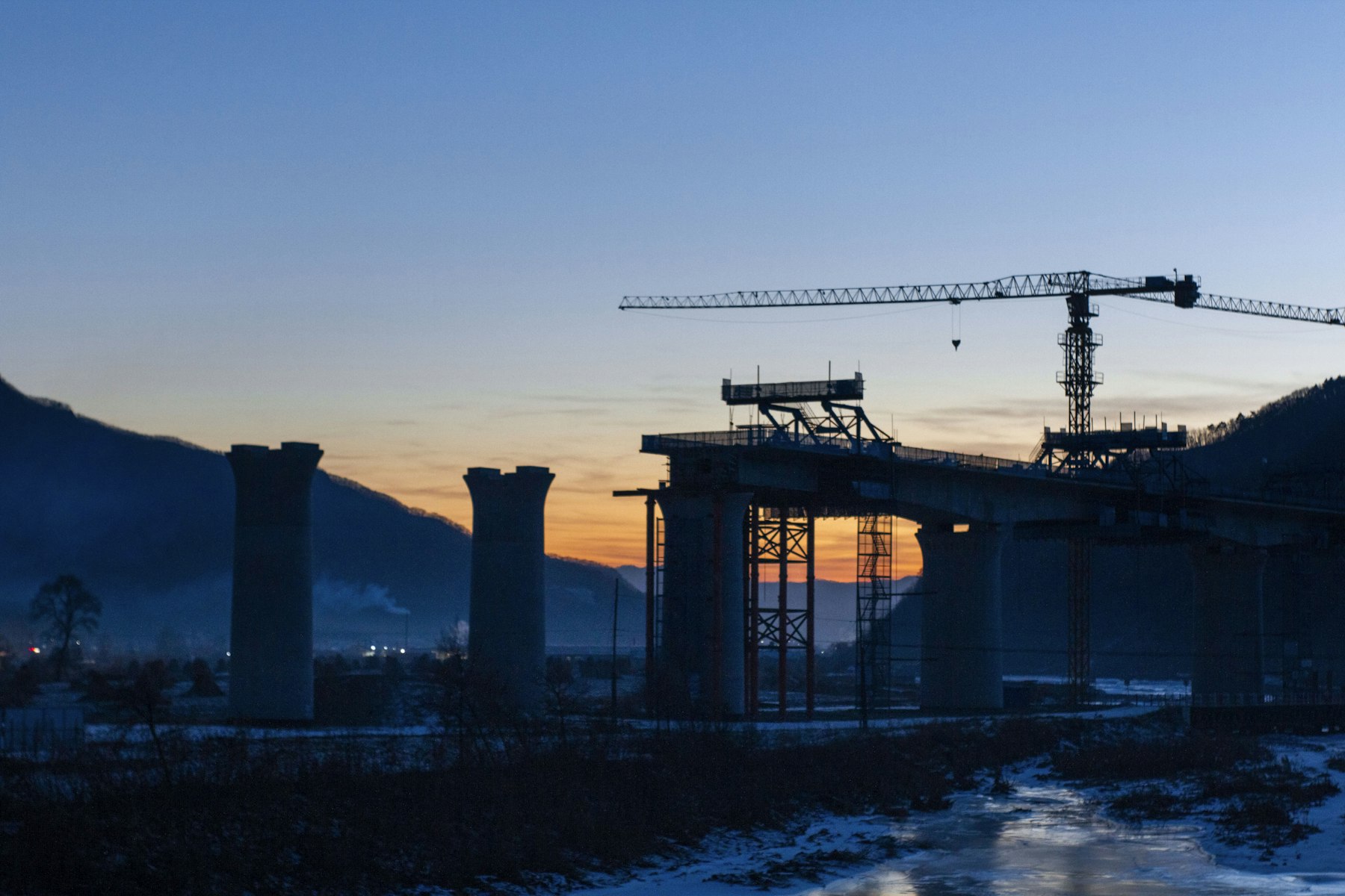 A tower crane and bridge piers under construction at dusk, with mountains in the background.
