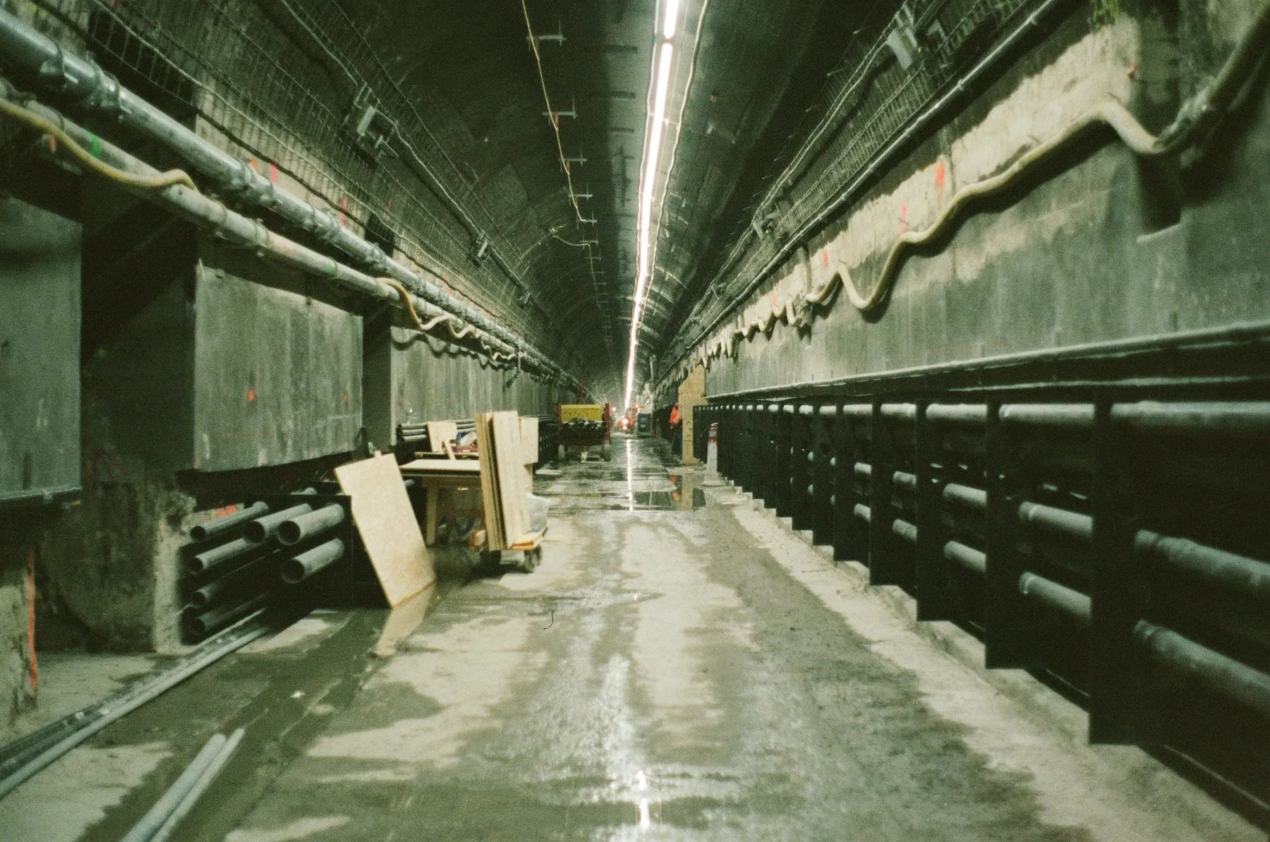 A long unfinished concrete tunnel lined with pipes, cables and lighting.