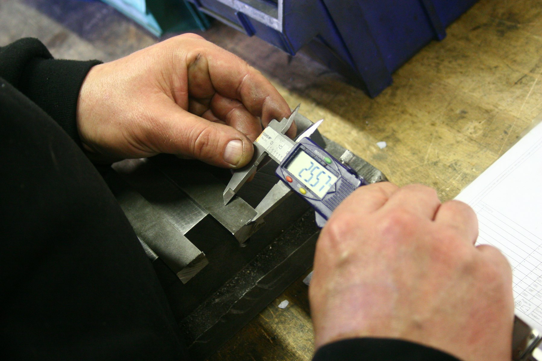 Hands using a digital caliper to measure a machined metal part.
