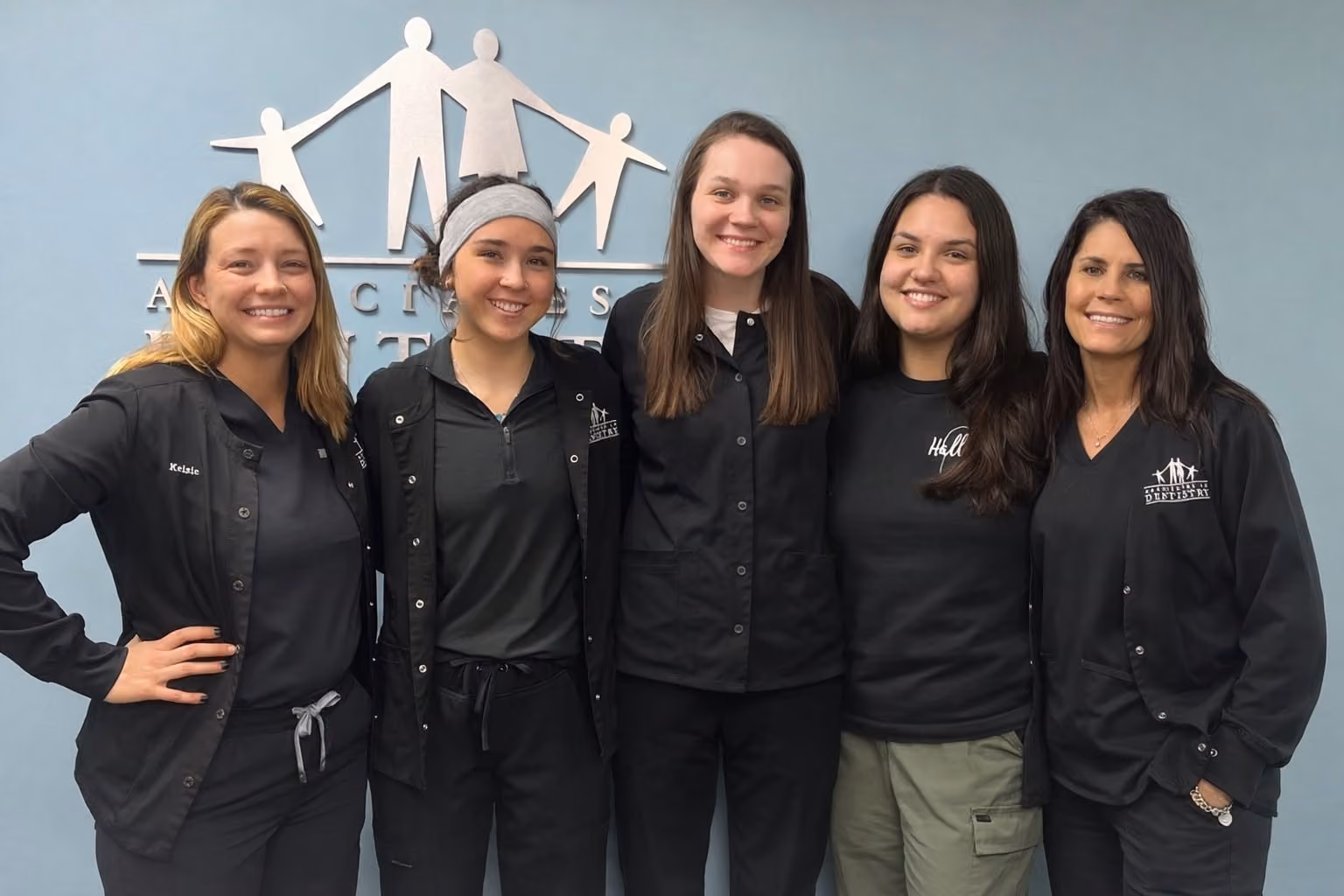 Group of five smiling women standing side by side in black medical or dental uniforms against a light blue wall with a white family-themed logo behind them.