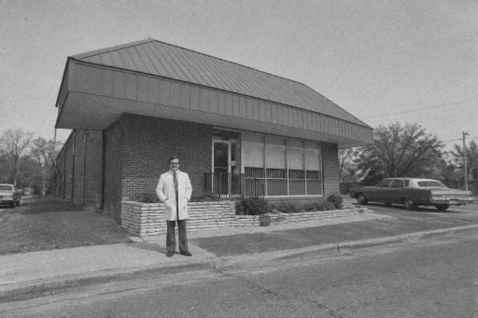 Black and white photo of a man in a white lab coat and tie standing on a sidewalk in front of a mid-century modern brick building with large windows and a metal roof; an old car is parked on the side street nearby.