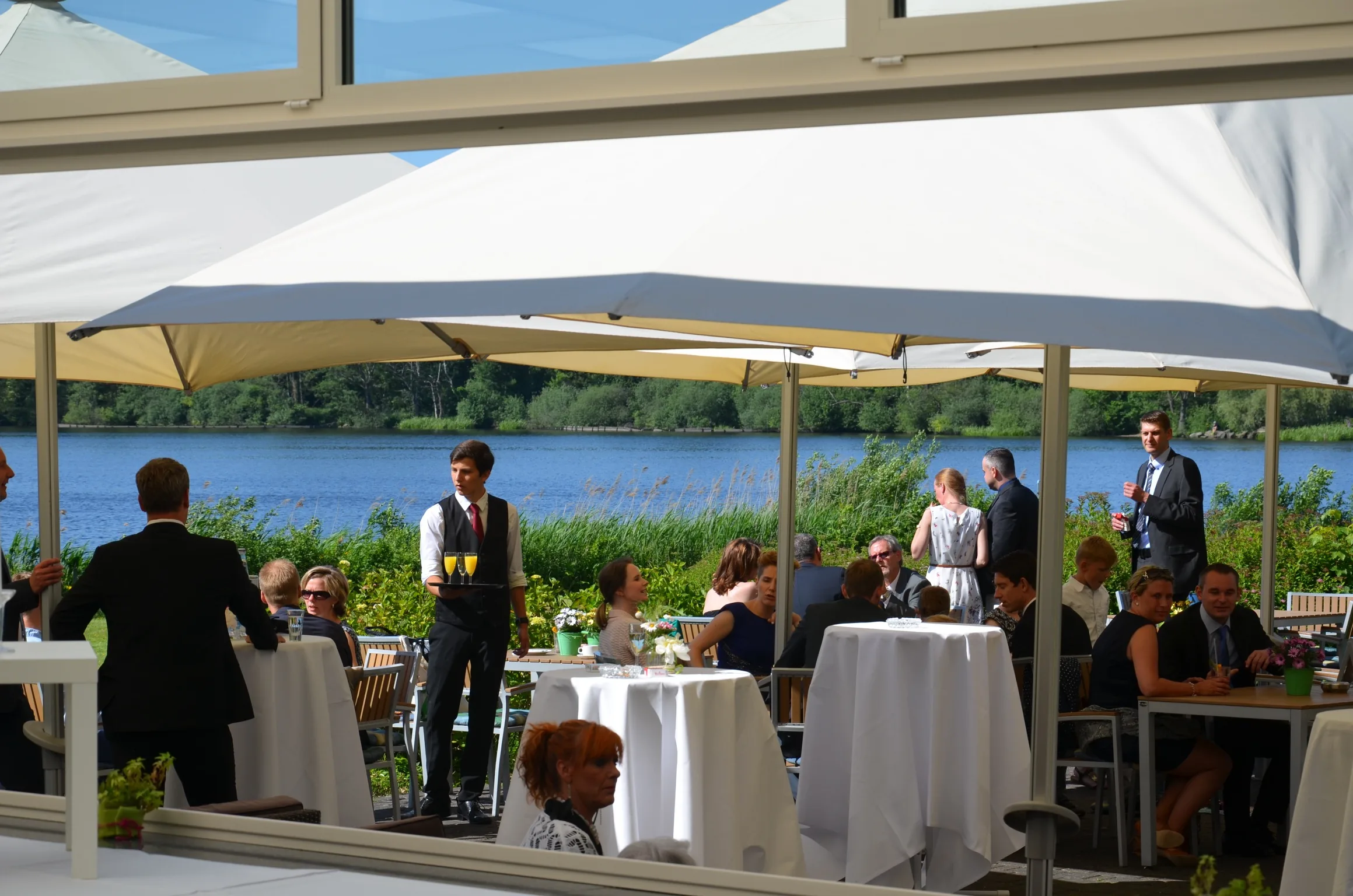 Gäste beim Sektempfang auf der Seeterrasse unter hellen Schirmen, dahinter der See