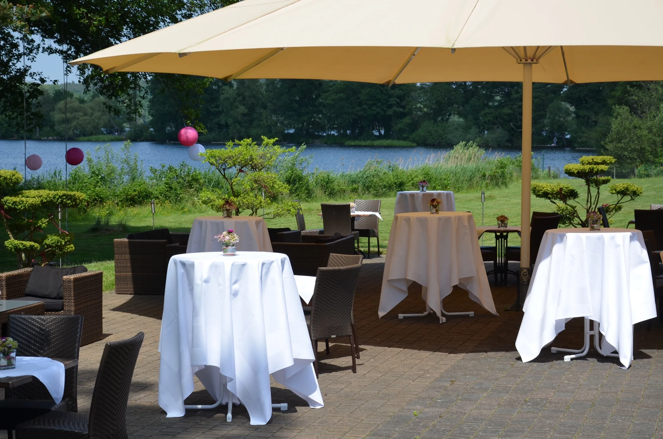 Stehtische mit weißen Hussen auf der Seeterrasse unter einem großen Schirm, dahinter der See