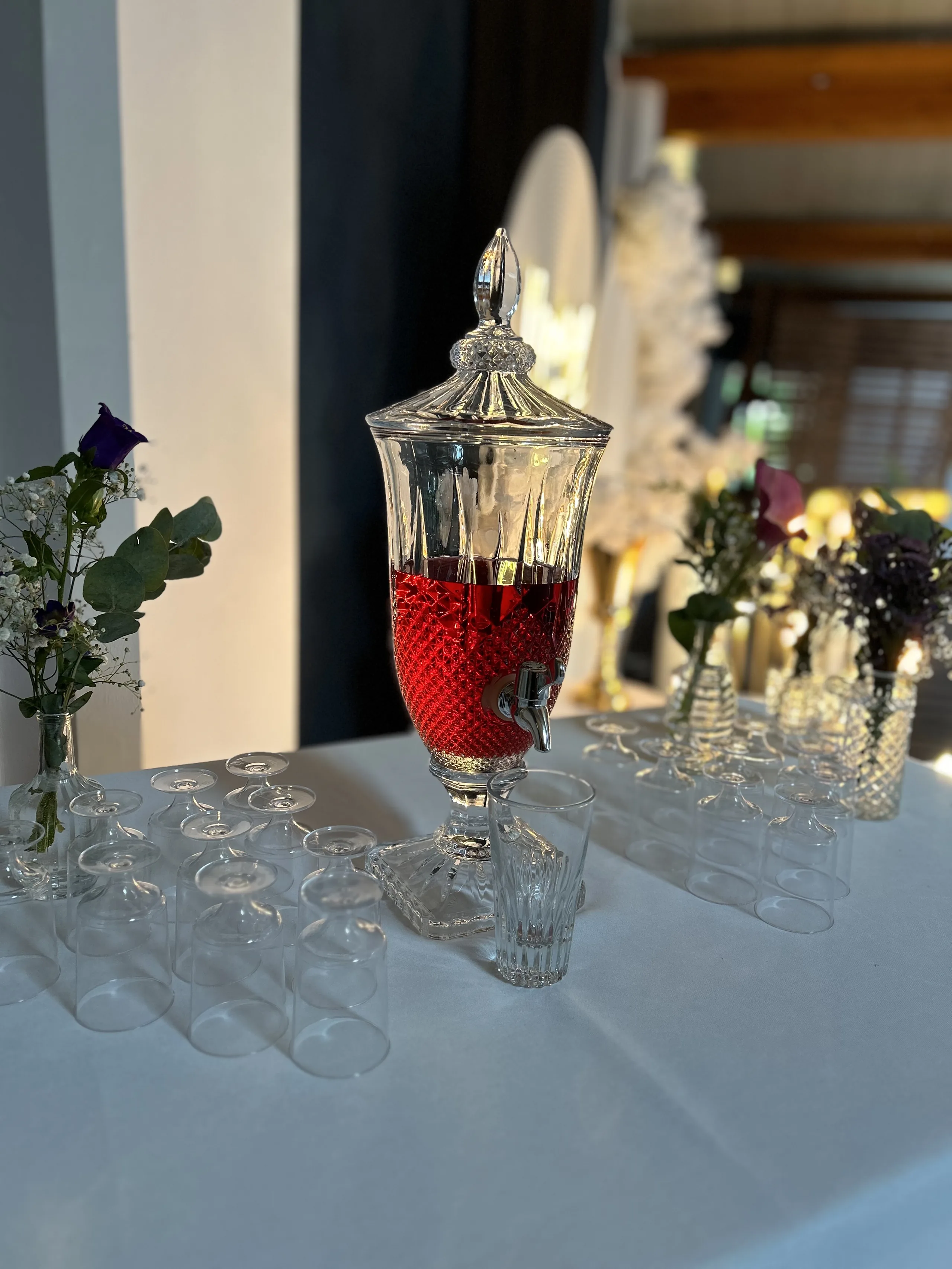 Getränkespender aus Glas mit rotem Tee, daneben Gläser und Blumen