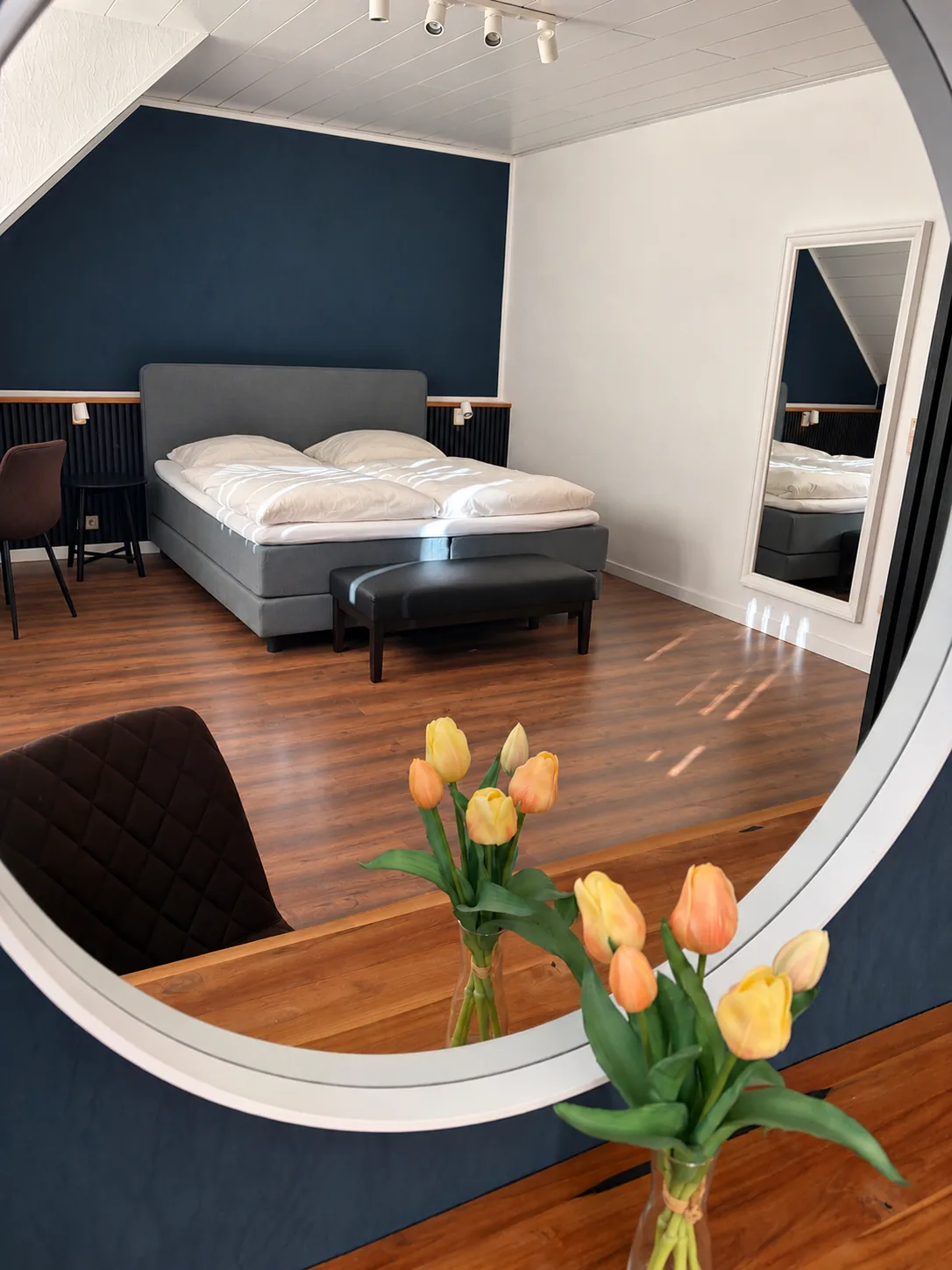 Blick durch einen runden Spiegel auf das Doppelzimmer mit Boxspringbett, vorne gelbe Tulpen
