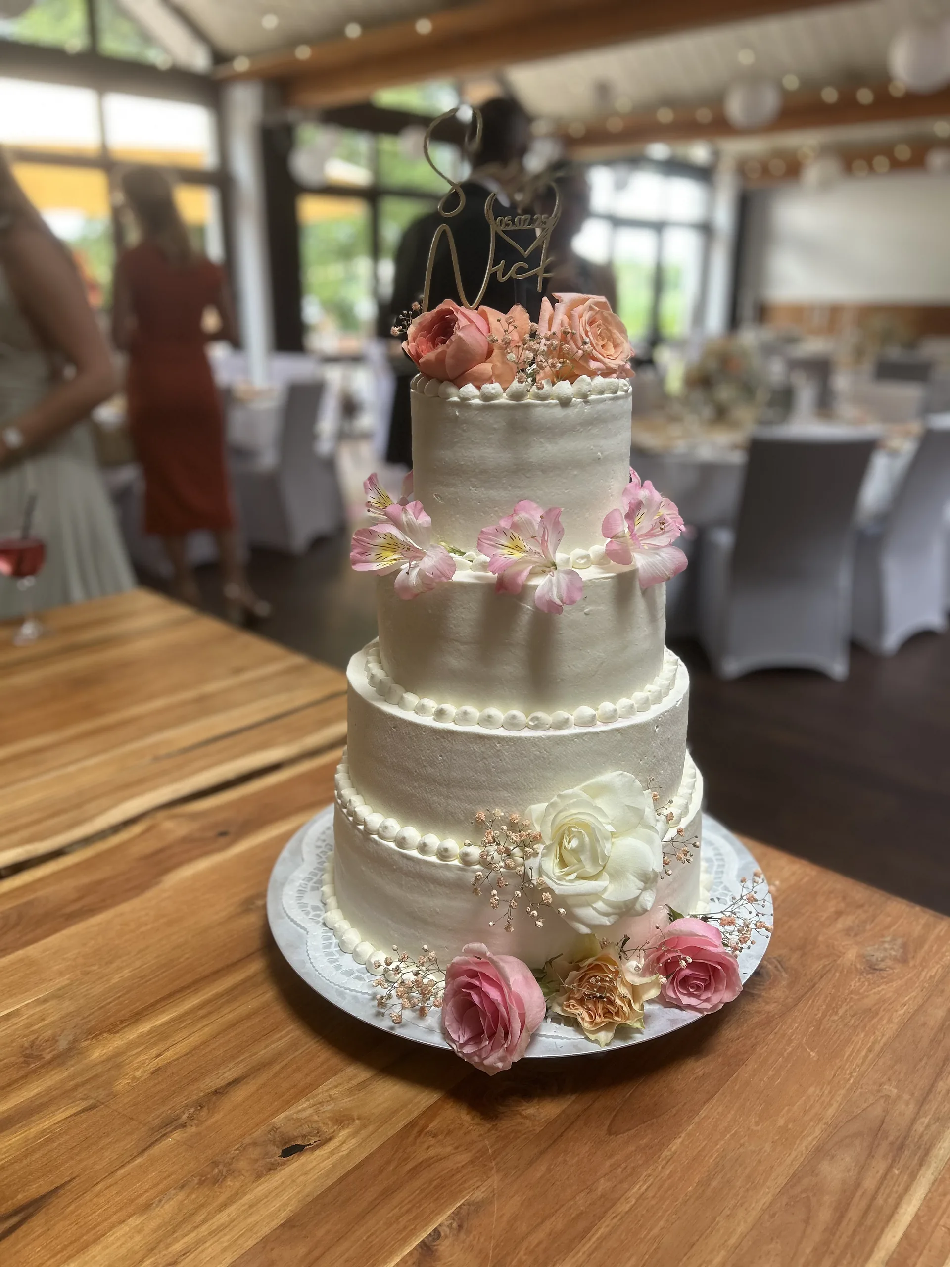 Dreistöckige Hochzeitstorte mit rosa Rosen, im Hintergrund der eingedeckte Saal