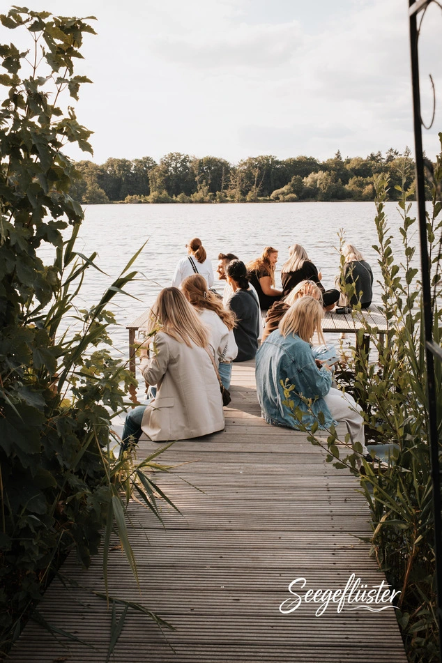 Gäste an Stehtischen auf der Seeterrasse unter hellen Schirmen, ein Kellner serviert, dahinter der See