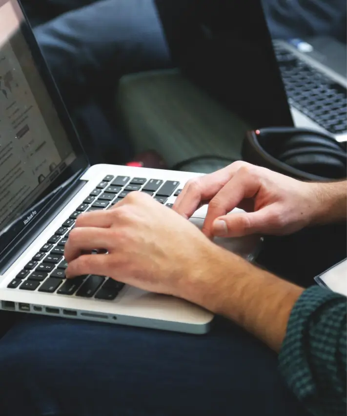 Person typing on a MacBook Pro laptop keyboard with headphones and another laptop in the background.