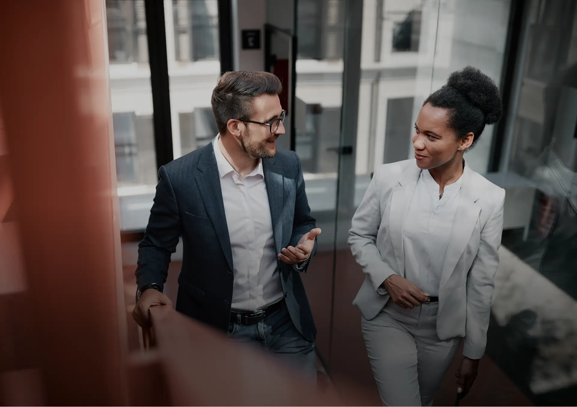 Two business professionals walking and talking in a modern glass office hallway.