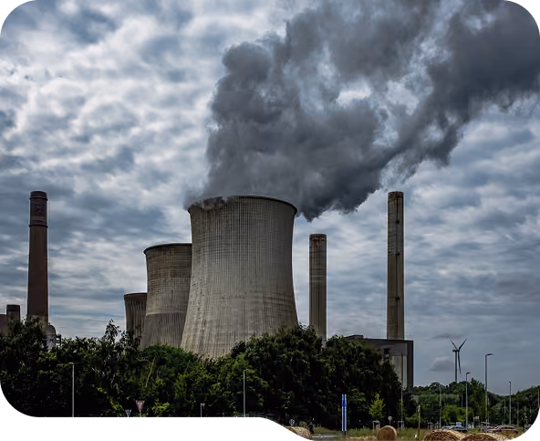 Industrial power plant with large cooling towers emitting thick dark smoke under a cloudy sky, surrounded by trees and road infrastructure.