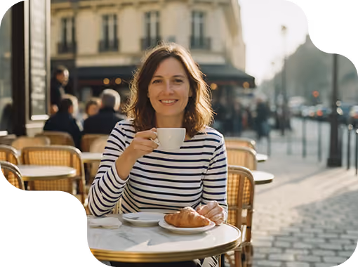 Smiling woman in a striped shirt sitting at an outdoor café table holding a white cup with a croissant on a plate in front of her.