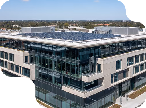 Modern multi-story office building with large glass windows and solar panels installed on the flat roof, surrounded by a suburban landscape under a clear blue sky.