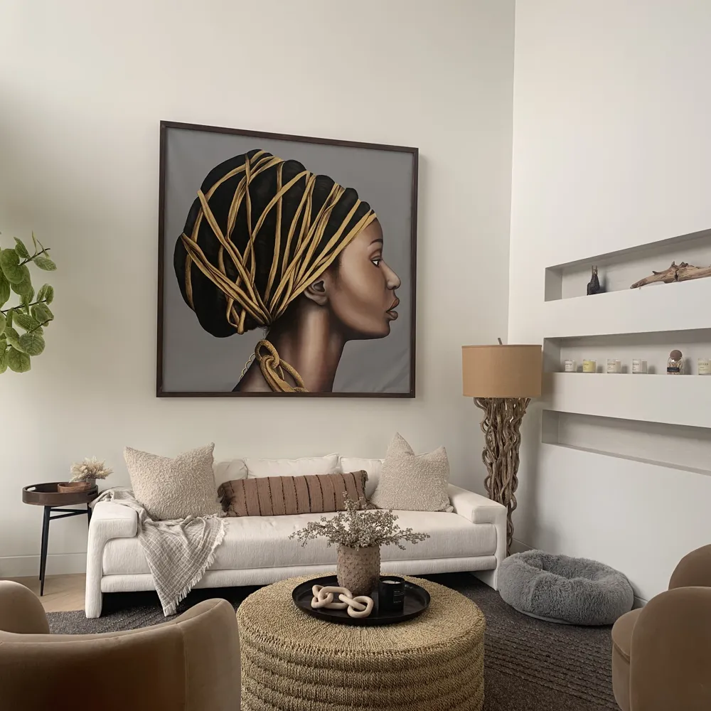 Modern living room with a white sofa adorned with beige and brown pillows, a cozy beige throw, a textured round woven coffee table with a vase and decorative items, a unique wooden floor lamp with a beige shade, a gray pet bed, and a large framed profile portrait of a woman with an intricate headwrap on the wall.
