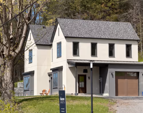 Modern two-story custom home with light-colored exterior, gray shingle roof, multiple black-framed windows, attached garage with wooden doors, and a small porch; surrounded by green grass and trees.