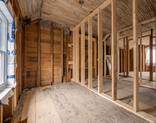 Interior of a house under renovation showing exposed wooden wall studs, ceiling joists, and a bare wooden floor with construction materials near a window on the left.