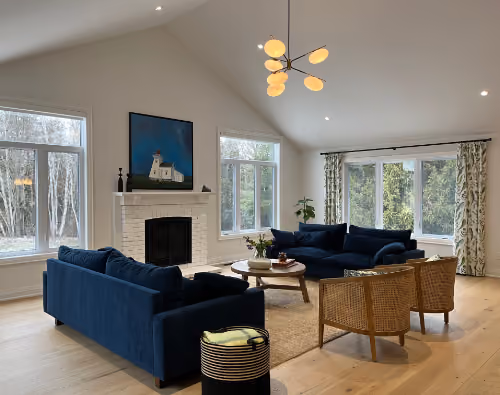 Modern living room with two blue sofas, two wicker chairs, a round wooden coffee table on a beige rug, large windows with natural light, white walls, a fireplace with artwork above it, and a contemporary ceiling light fixture.