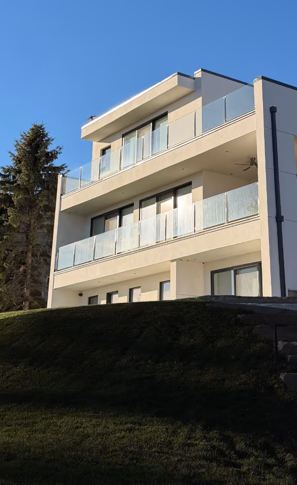 Modern three-story white building with large glass balcony railings, situated on a grassy hill with a tree to the side and clear blue sky.