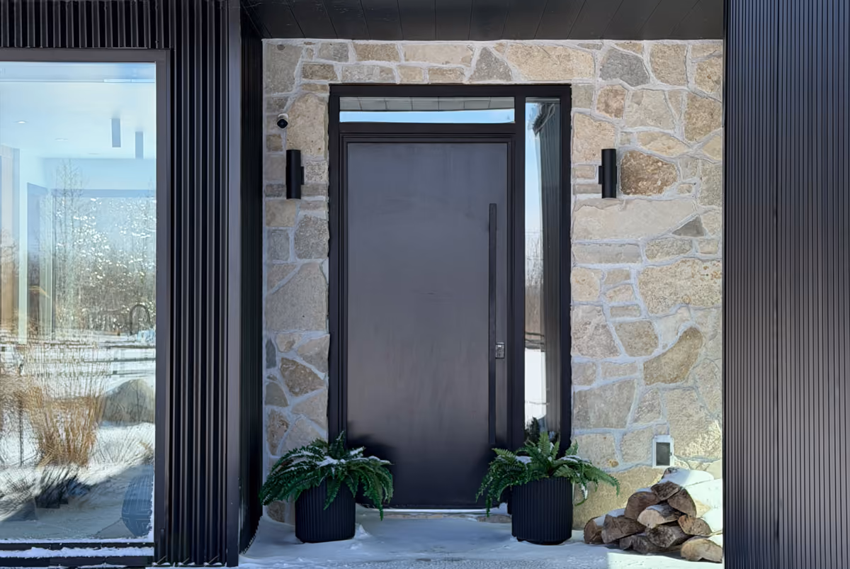 Modern black front door with large vertical handle set in a stone wall, flanked by two black wall lights, two black plant pots with green ferns, and a stack of firewood on a snowy porch.