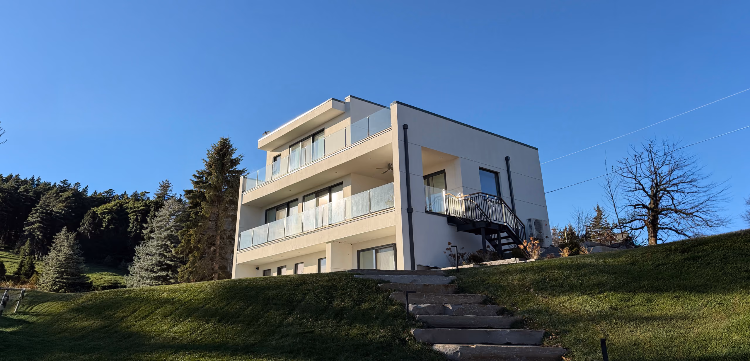 Modern two-story white house with large glass balconies situated on a grassy hill under a clear blue sky.