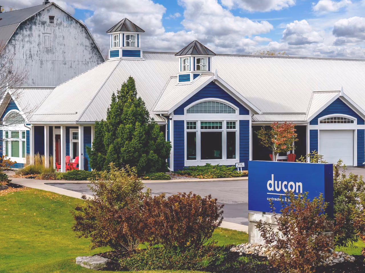 Blue and white building with a metal roof and two cupolas in front of a partly cloudy sky, surrounded by greenery and bushes, with a blue sign reading 'ducon'.