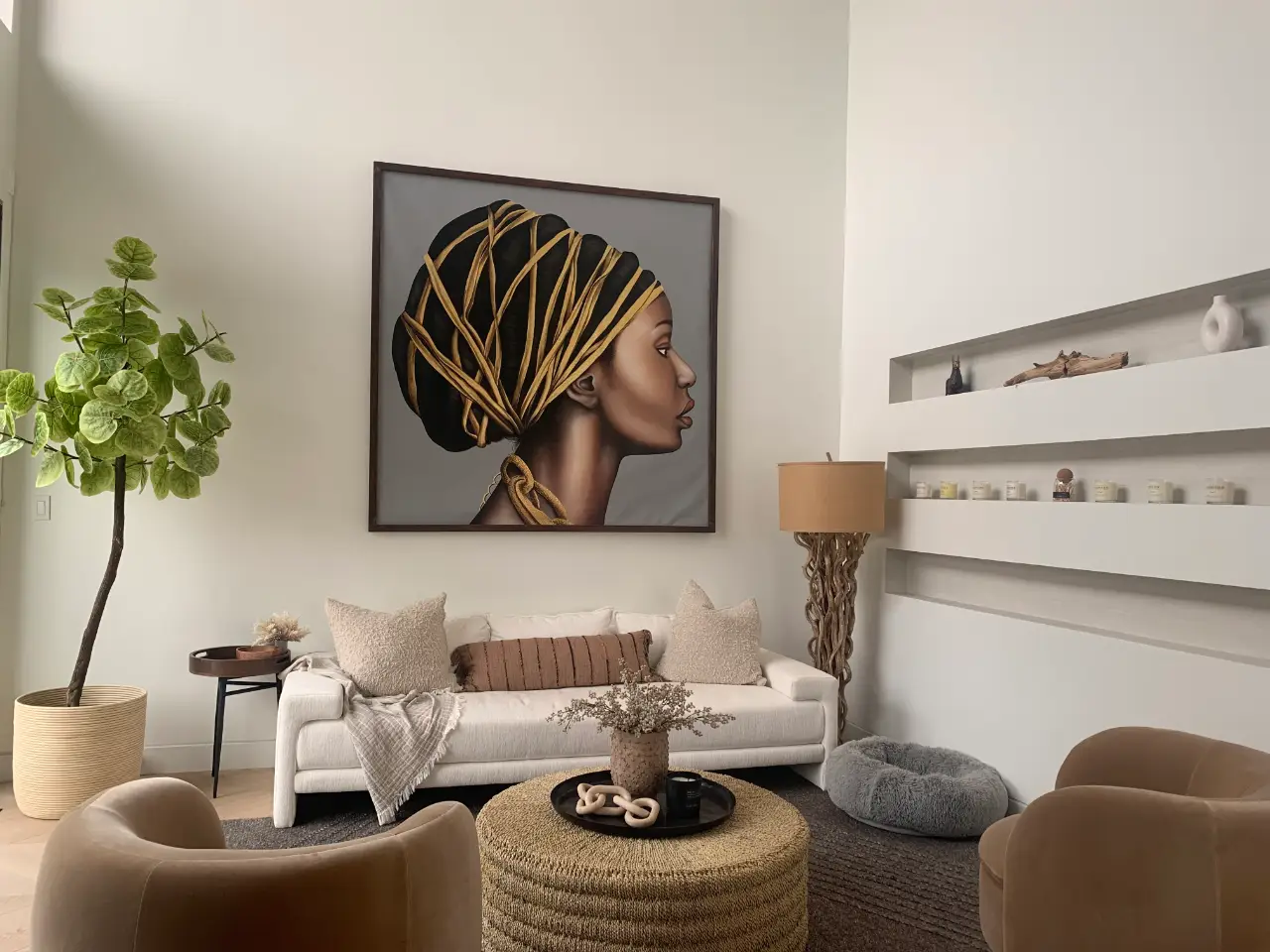 Modern living room with a white sofa adorned with beige and brown pillows, a cozy beige throw, a textured round woven coffee table with a vase and decorative items, a unique wooden floor lamp with a beige shade, a gray pet bed, and a large framed profile portrait of a woman with an intricate headwrap on the wall.