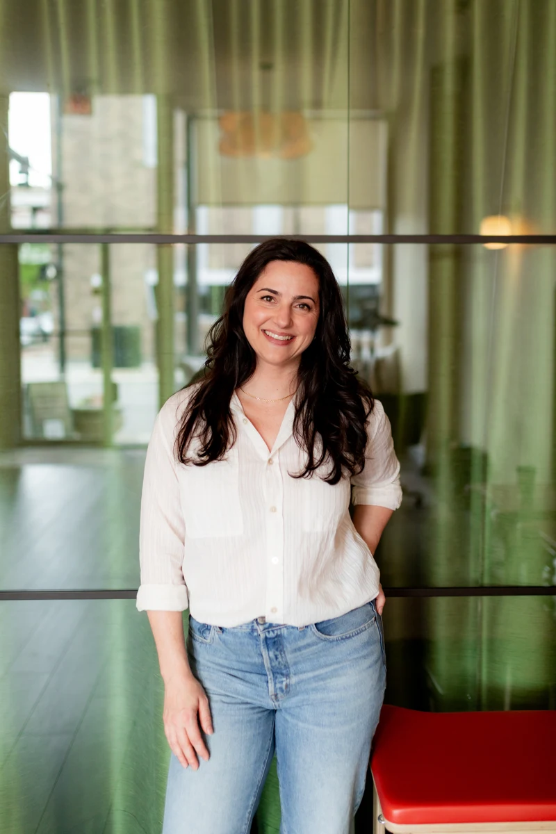 Holly Swanson smiling, standing in front of a glass wall in a white shirt and jeans.